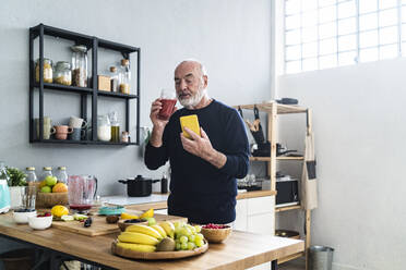 Älterer Mann mit Smartphone trinkt Smoothie zu Hause - GIOF13833