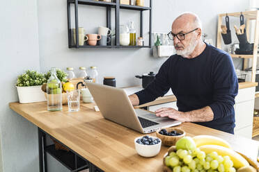 Älterer Mann, der an der Küchentheke sitzend einen Laptop benutzt - GIOF13834