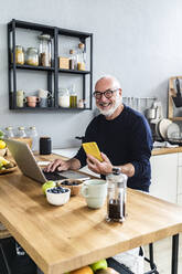 Smiling senior man with mobile phone and laptop sitting at kitchen counter - GIOF13842