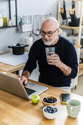 Älterer Mann trinkt Wasser, während er zu Hause einen Laptop benutzt - GIOF13844