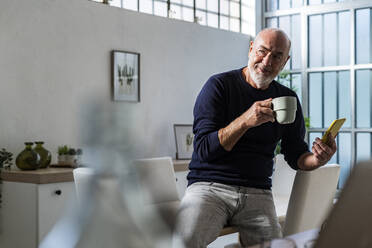 Älterer Mann mit Kaffeetasse und Smartphone auf dem Tisch sitzend zu Hause - GIOF13852