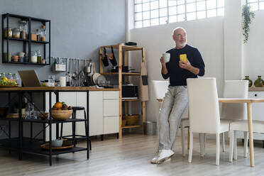 Senior man with mobile phone and coffee cup leaning on dining table - GIOF13855