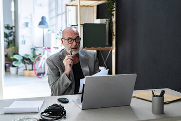 Männlicher Berufstätiger mit Dokumenten und Laptop an einem Schreibtisch im Studio sitzend - GIOF13861