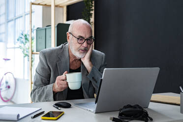 Geschäftsmann mit Kaffeetasse und Blick auf den Laptop im Studio - GIOF13863
