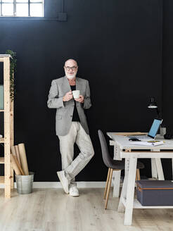 Älterer Geschäftsmann mit Kaffeetasse an schwarzer Wand lehnend im Studio - GIOF13866