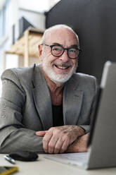Smiling senior male professional sitting with laptop in studio - GIOF13872