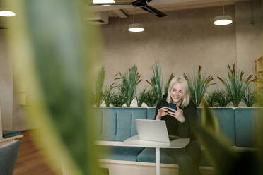 Lächelnde Geschäftsfrau, die ein Smartphone benutzt, während sie in einem Coworking-Büro sitzt - OGF01093