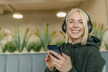 Fröhliche blonde Geschäftsfrau hört Musik über drahtlose Kopfhörer in einem Coworking-Büro - OGF01098