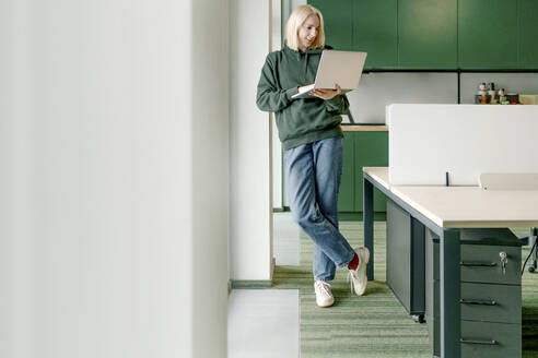 Businesswoman using laptop while leaning on architectural column in office - OGF01103