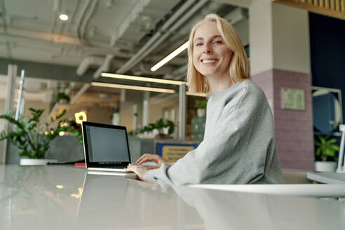 Smiling businesswoman using laptop on table in coworking office - OGF01107