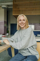 Blond businesswoman with laptop sitting at table in coworking office - OGF01109