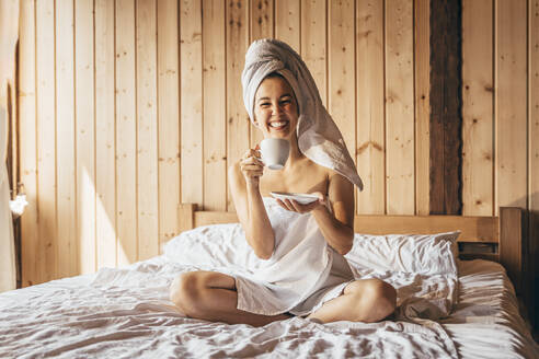 Glückliche Frau im Handtuch, die Kaffee trinkt, während sie auf dem Bett sitzt - OMIF00142