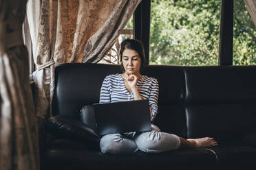 Frau mit Hand auf dem Kinn bei der Benutzung eines Laptops auf der Couch zu Hause - OMIF00148