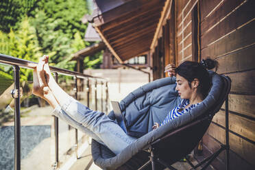 Woman using laptop while sitting on chair in balcony - OMIF00151