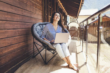 Smiling woman looking up while sitting on chair with laptop in balcony - OMIF00153
