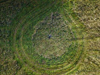 Aerial view of lone woman lying in green field encircled by tire tracks - KNTF06497