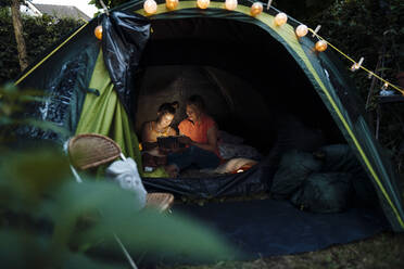 Grandmother and granddaughter camping in garden using digital tablet in tent - MOEF03953
