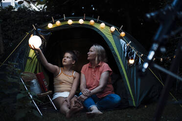 Girl holding illuminated lantern while sitting with grandmother at backyard - MOEF03956