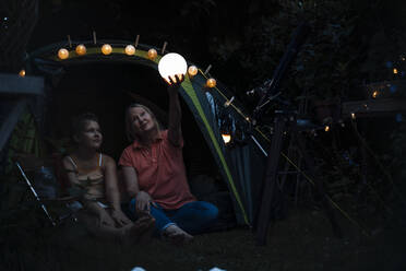 Grandmother holding illuminated lantern while sitting with girl at tent entrance - MOEF03957