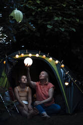 Senior woman holding illuminated lantern while sitting with girl at tent entrance - MOEF03959