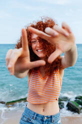 Positive Frau mit langem lockigem rotem Haar, die ein Dreieck am Meeresufer mit Felsbrocken formt und in die Kamera schaut - ADSF31140