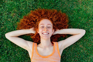 Top view of happy curly haired female with hands behind head relaxing on lawn and looking at camera - ADSF31142