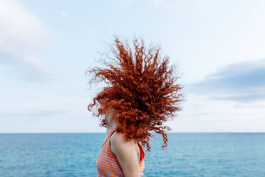 Seitenansicht einer unbekümmerten, anonymen Frau, die ihr lockiges, rotes Haar an der Küste des blauen Meeres schüttelt - ADSF31151