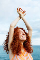 Delighted female with curly long ginger hair wearing knitted sweater standing with arms raised and closed eyes against blue sea - ADSF31155