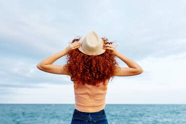 Back view of unrecognizable female with long ginger curls with hat standing on coast of blue sea - ADSF31156