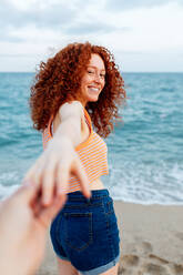 Back view of carefree young female traveler with curly ginger hair holding hand of crop boyfriend on sandy beach washed by blue sea - ADSF31159