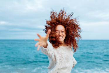 Pleasant female with long ginger curly hairstyle reaching hand to camera while standing against waving sea - ADSF31160