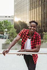 Fröhlicher afroamerikanischer Mann in kariertem Hemd, der sich an ein Metallgeländer lehnt und in einem Stadtpark fröhlich lacht - ADSF31165