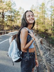 Seitenansicht einer glücklichen weiblichen Reisenden mit Rucksack, die in die Kamera schaut, während sie auf einer asphaltierten Straße auf Teneriffa (Kanarische Inseln, Spanien) geht - ADSF31173
