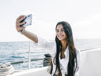 Lächelnder weiblicher Teenager mit langen Haaren, der ein Selbstporträt mit dem Handy auf einem vertäuten Motorboot auf dem Meer in Teneriffa Spanien aufnimmt - ADSF31176