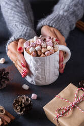 From above of crop female holding mug of hot drink with marshmallows among Christmas gifts cones and cinnamon sticks - ADSF31184