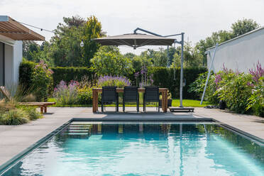 Esstisch mit Stuhl und hölzernen Sonnenliegen in der Nähe des Swimmingpools im Hof einer teuren, modernen, minimalistisch gestalteten Villa an einem sonnigen Tag - ADSF31187