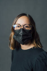 Young woman wearing black protective face mask against gray background - MEUF04437