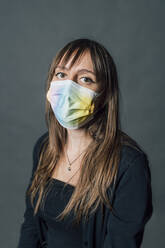 Young woman wearing multi colored protective face mask against gray background - MEUF04447