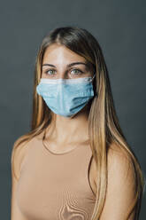 Young woman with long hair wearing protective face mask against gray background - MEUF04495