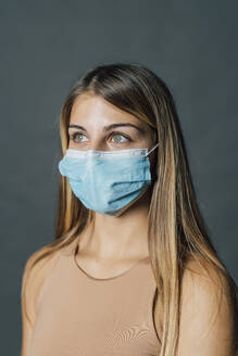 Woman with long hair wearing protective face mask against gray background - MEUF04496