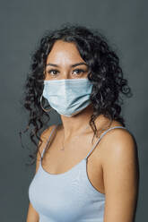 Woman with curly hair wearing protective face mask against gray background - MEUF04501