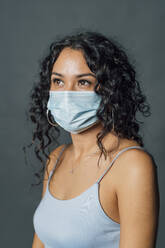 Woman with black curly hair wearing protective face mask against gray background - MEUF04502