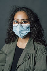 Young woman with eyeglasses wearing protective face mask against gray background - MEUF04508
