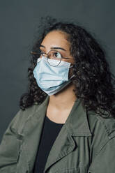 Thoughtful woman with eyeglasses wearing protective face mask against gray background - MEUF04509