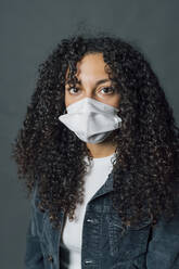 Woman with curly hair wearing protective face mask against gray background - MEUF04539