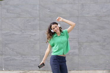 Carefree woman with wireless headphones dancing in front of wall - EIF02186