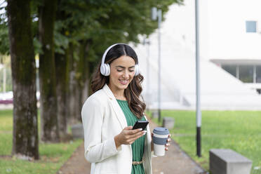 Happy woman with wireless headphones using mobile phone at park - EIF02190