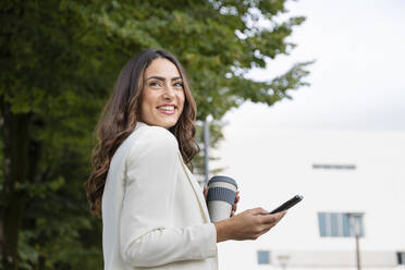 Lächelnde junge Frau mit Mobiltelefon und Einweg-Kaffeebecher im Park - EIF02194