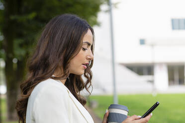 Woman holding disposable cup using mobile phone at park - EIF02195