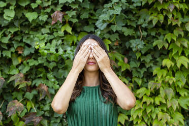 Young woman covering eyes with hands in front of green ivy plants - EIF02215